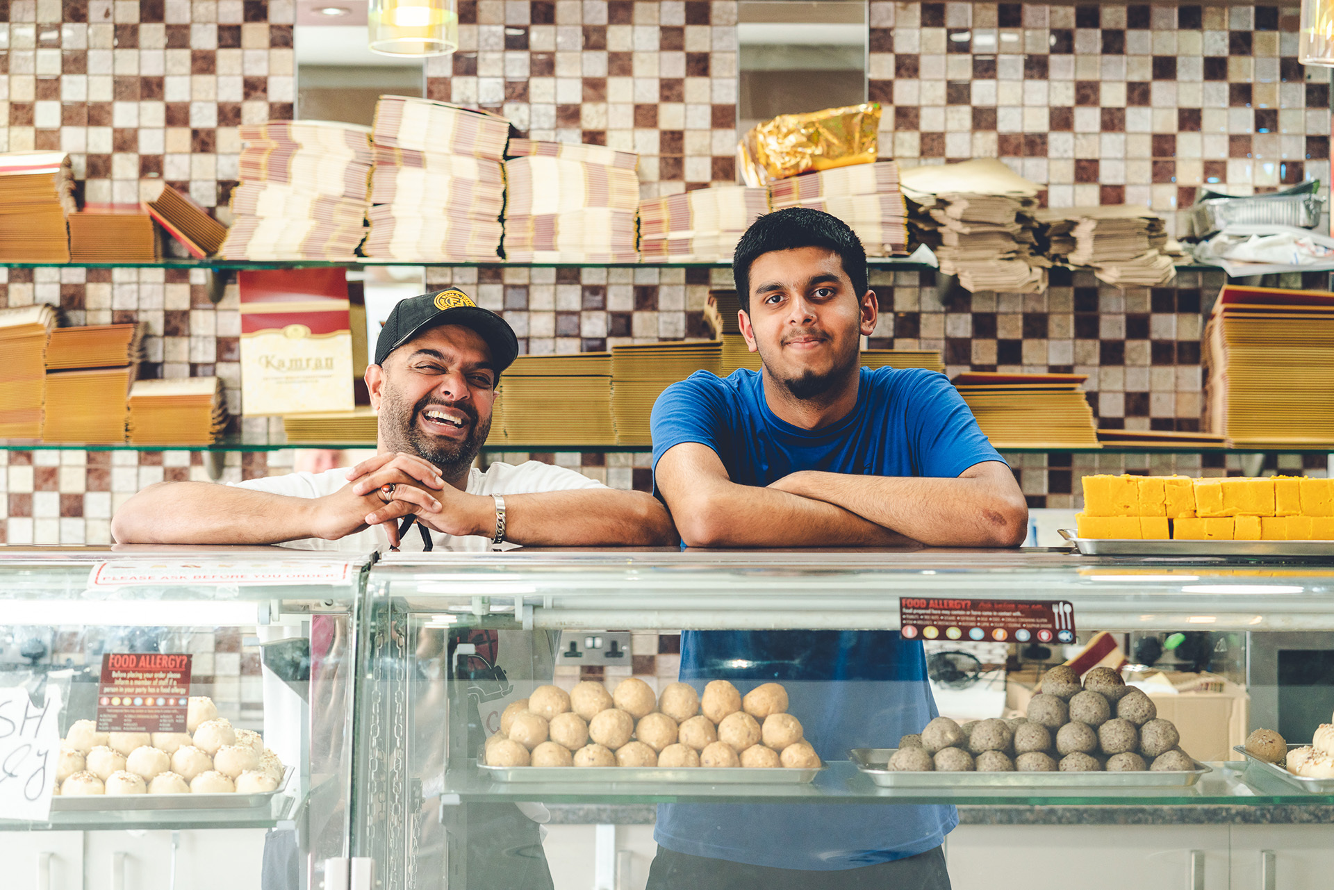 Photograph of two men standing behind the counter of Kamran Sweet Centre. They have there arms folded on the countertop and are smiling at the camera. Inside the counter are lots of delicious-looking sweet treats.