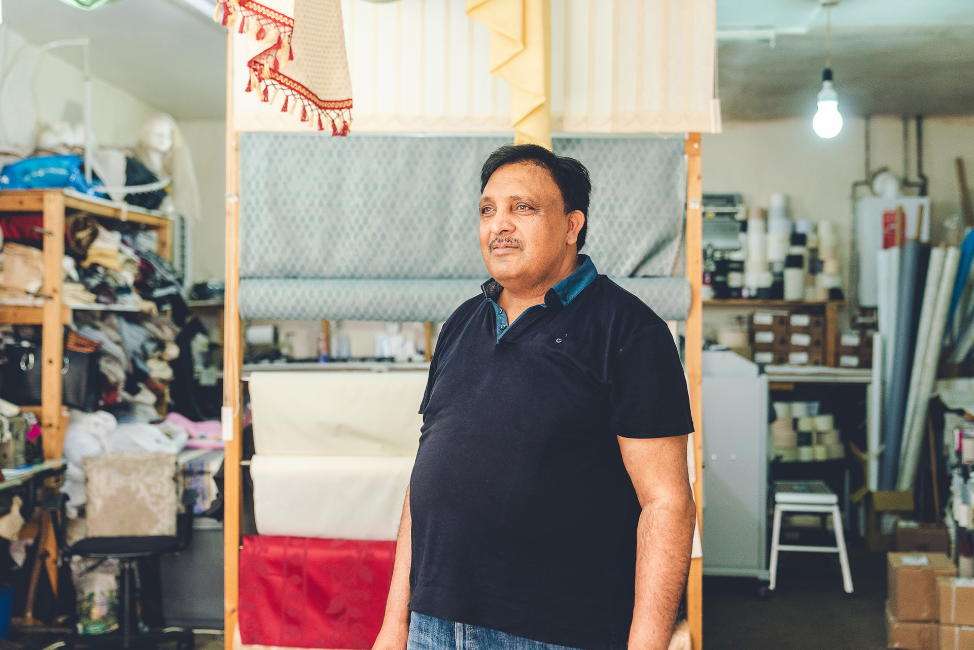 Photograph of a man standing inside a fabric store. Around him are folds and rolls of fabric of different colours and patterns.