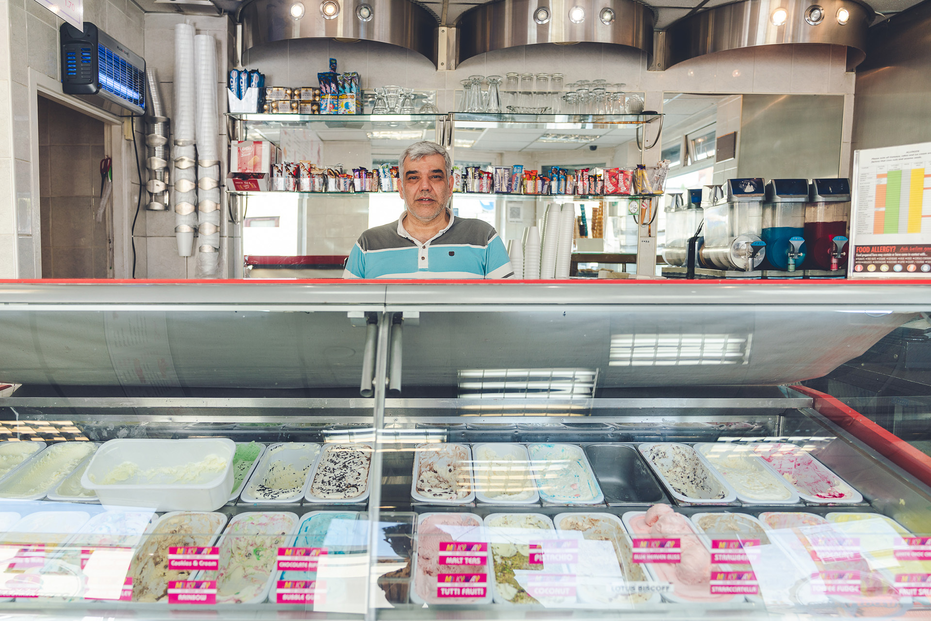 Photograph of a men standing behind the counter in Milky Way. The counter is filled with lots of different flavours of ice cream.