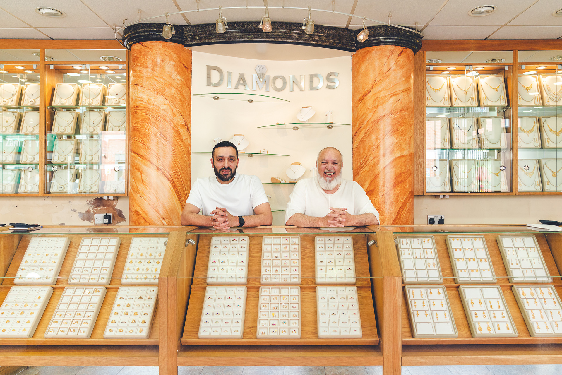 Photograph of two men inside Naz Jewellers. Both stand behind the jewellery counter, smiling towards the camera. Around them are impressive displays of jewellery.