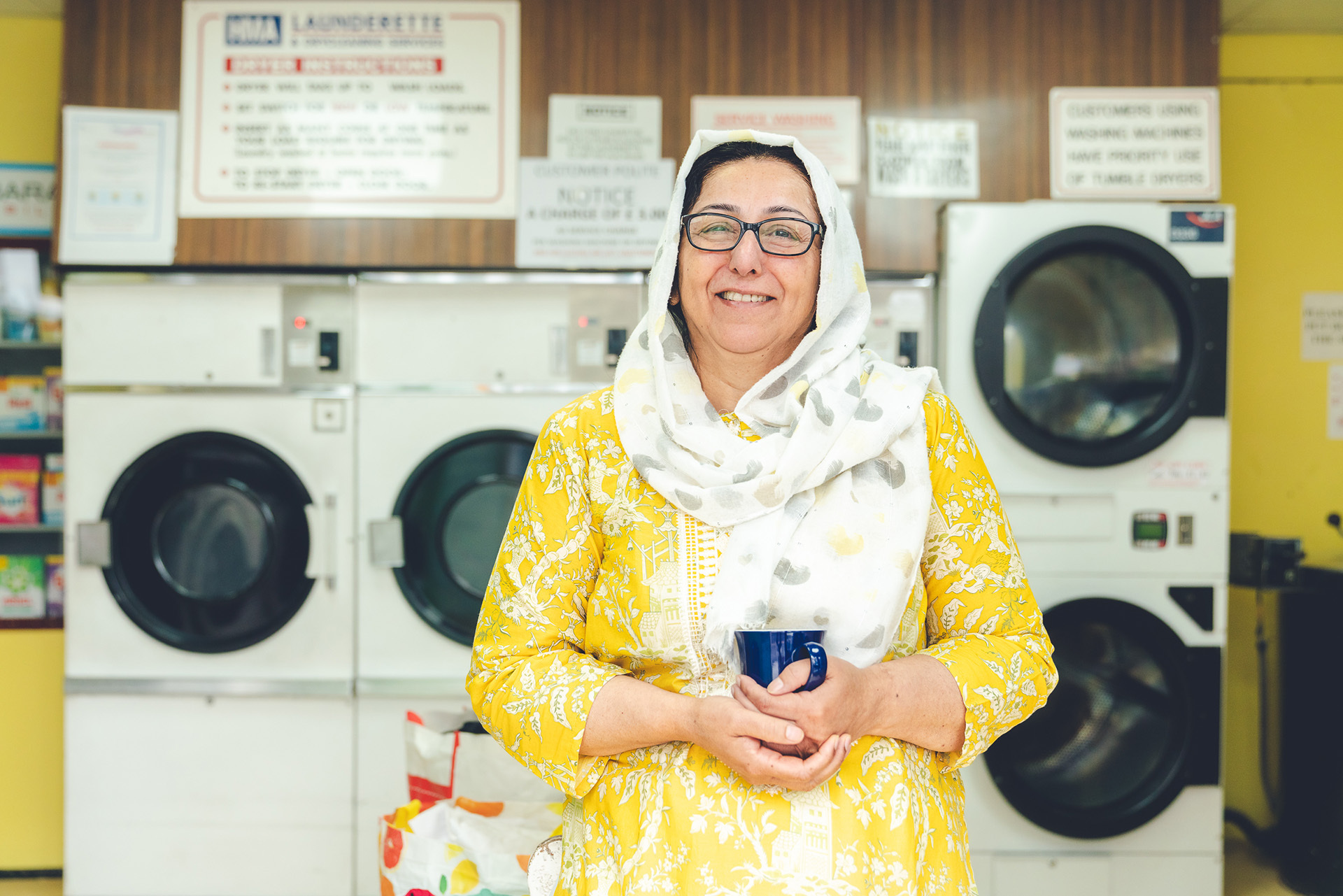 Photograph of a woman inside Virgo Laundrette, she is brightly dressed in yellow with a white headscarf. She holds a cup of tea and smiles at the camera.