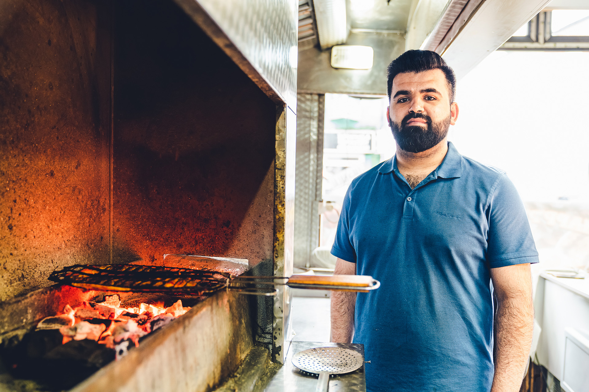 Photograph of a man standing next to a flame grilled inside Al Qudds. He looks proudly towards the camera.