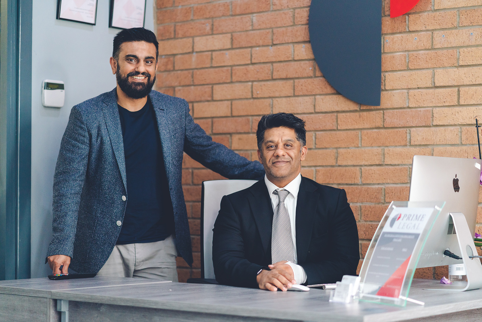 Photograph of two men from Prime Solicitors. One man is seated at a desk, the other stands to his right with his hand on the chair. Both are smiling.