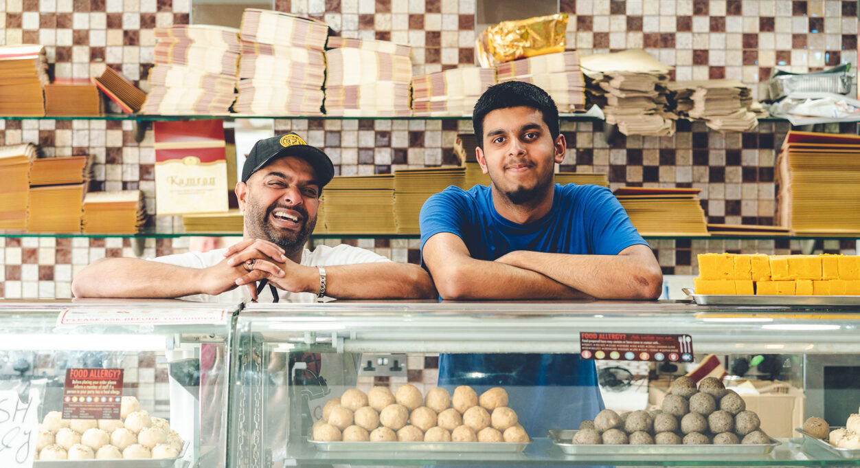 Photograph of two men standing behind the counter of Kamran Sweet Centre. They have there arms folded on the countertop and are smiling at the camera. Inside the counter are lots of delicious-looking sweet treats.